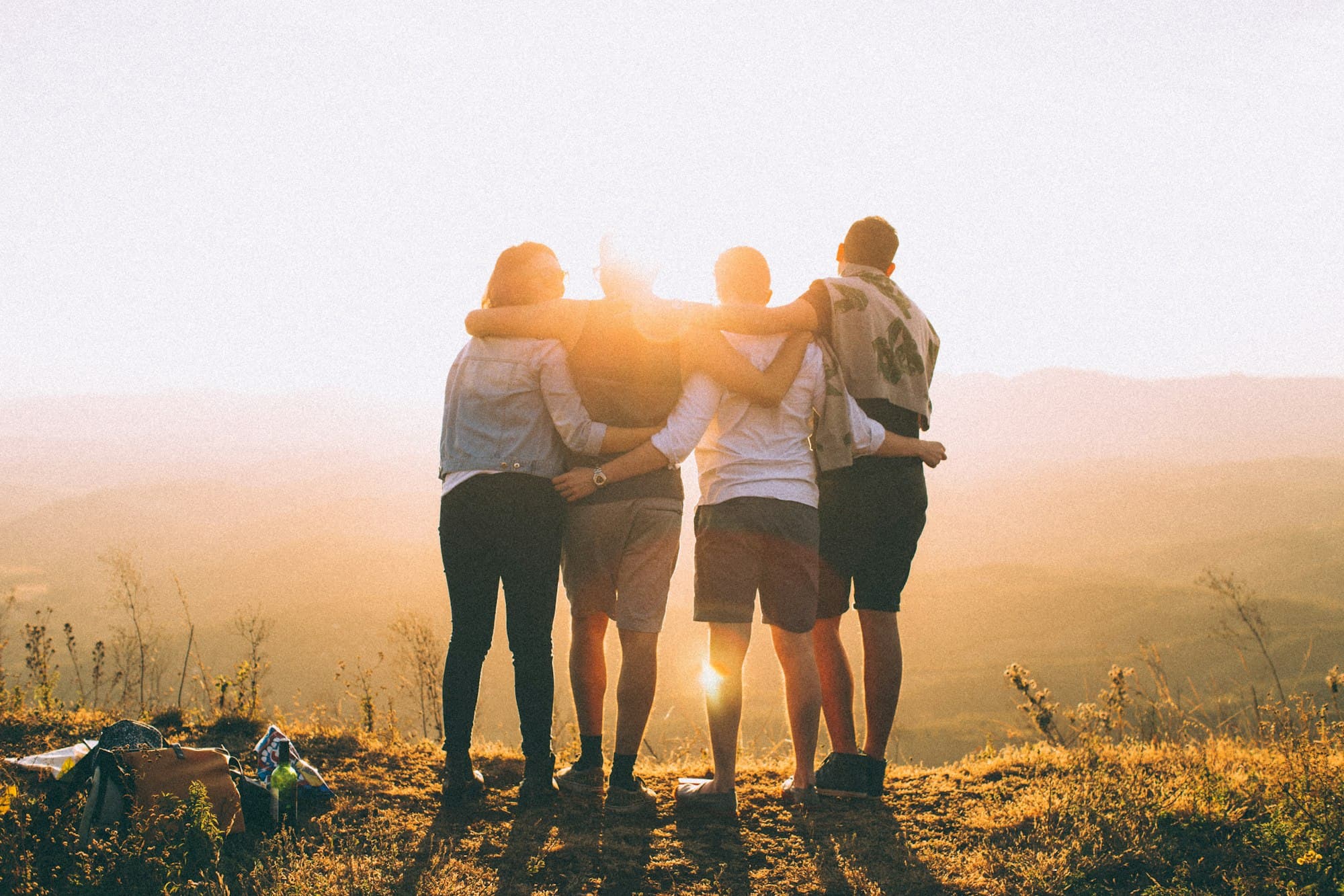 Four people standing close together on a grassy hill at sunset, with their arms around one another.